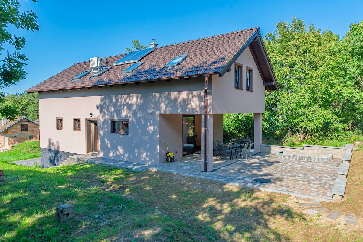 A spacious modern residence is depicted with a stone patio and wooden accents. Large windows provide ample natural light, and a shaded area includes a table and chairs for outdoor gatherings. Lush greenery surrounds the property, creating a serene outdoor environment.