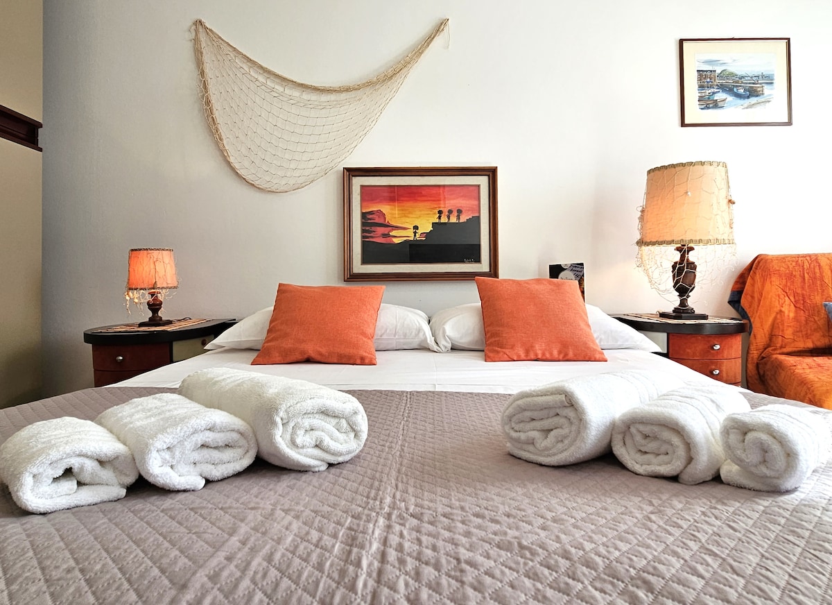 A spacious bedroom is presented with a large bed adorned with white pillows and vibrant orange cushions. Fluffy towels are neatly rolled and placed on the bed. Soft lamps illuminate the room, and artwork is displayed on the wall, contributing to the welcoming ambiance.