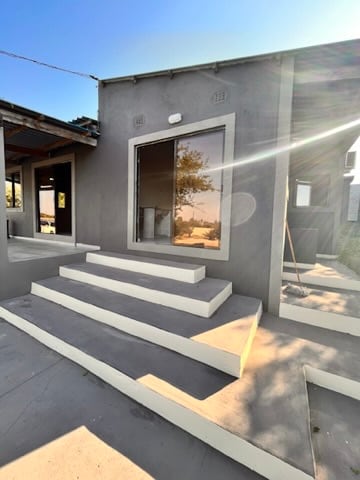 The exterior view shows a modern building with a minimalist design, featuring steps leading to a large window. Sunlight reflects off the glass, enhancing the clean lines of the structure. Surrounding greenery is visible in the background, suggesting a connection to nature.