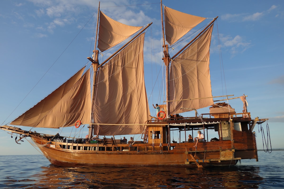 A traditional wooden ship with large, tan sails is anchored on calm waters. The boat features a sturdy wooden structure with visible deck rails and crew members preparing activities. Soft blue skies and scattered clouds add to the serene backdrop, highlighting a peaceful maritime atmosphere.