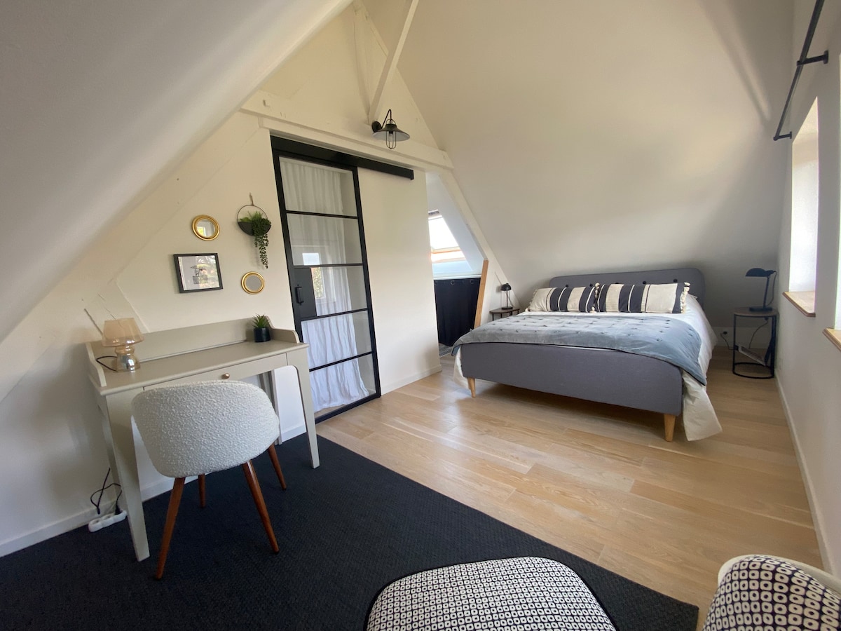 An attic bedroom features a spacious bed dressed in soft linens, accompanied by a cozy work desk. Natural light streams through a window, creating a bright and airy atmosphere. A sliding glass door adds a modern touch, while tasteful decor elements enhance the serene environment.