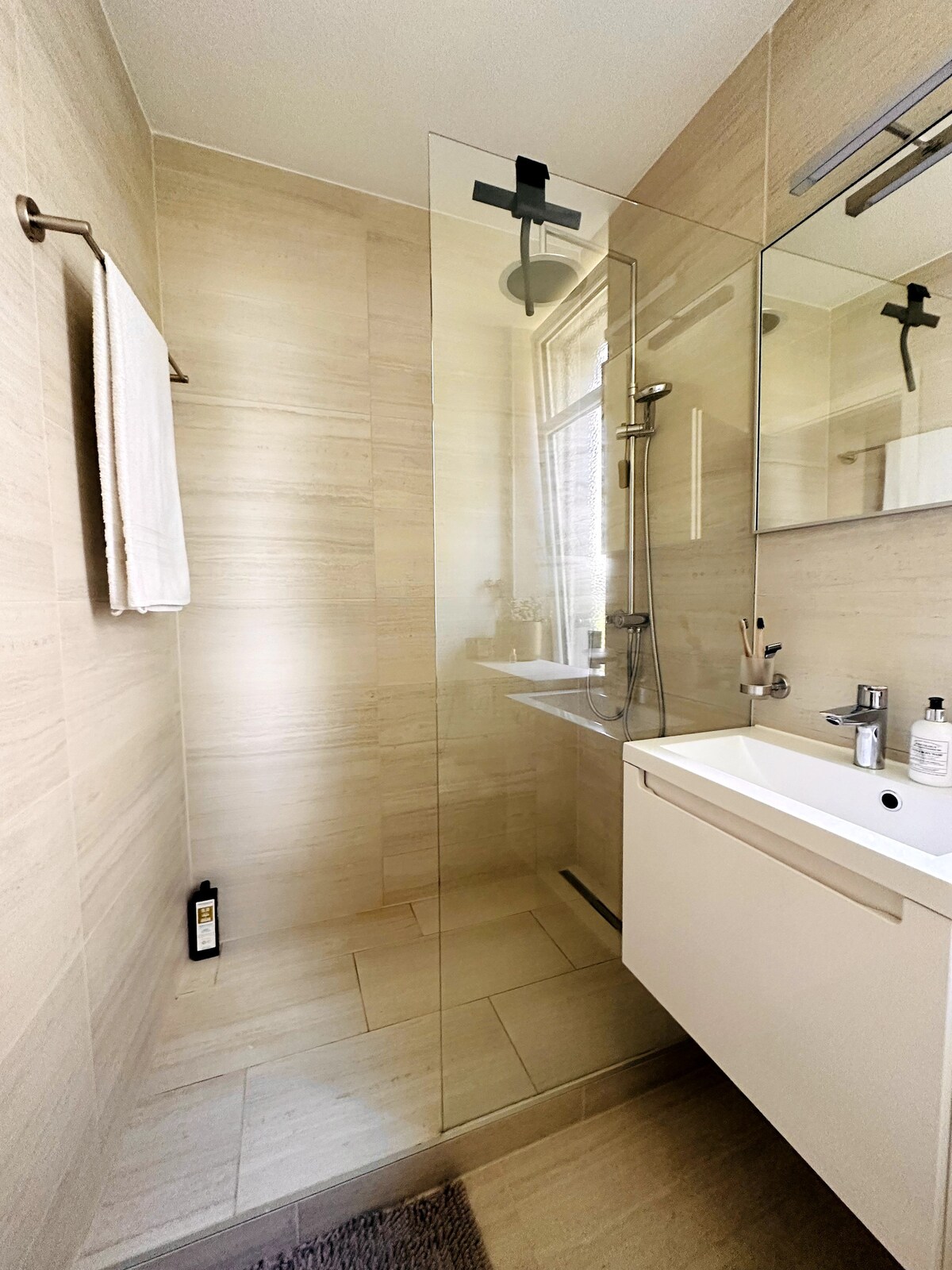 A modern bathroom features a glass-enclosed shower with a rainfall showerhead and a sleek vanity. Neutral-toned tiles create a calming atmosphere. A large mirror enhances the sense of space, while essential toiletries are neatly arranged on the counter.