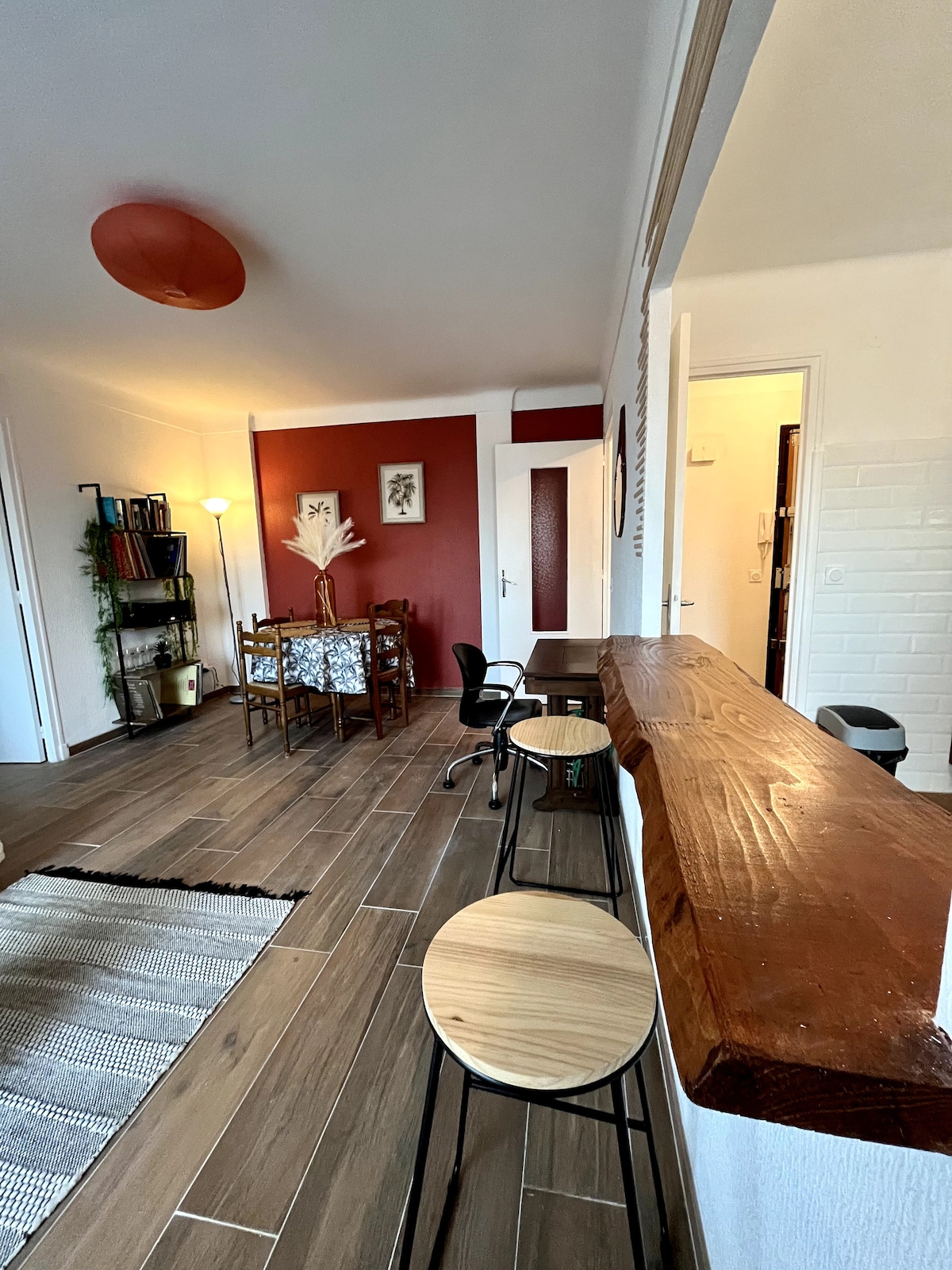 The living area features a warm colour palette, with a dining table for four and two stylish chairs. A bar counter separates the space, while wooden stools provide additional seating. Soft lighting creates a welcoming ambiance, enhancing the open layout of the apartment.