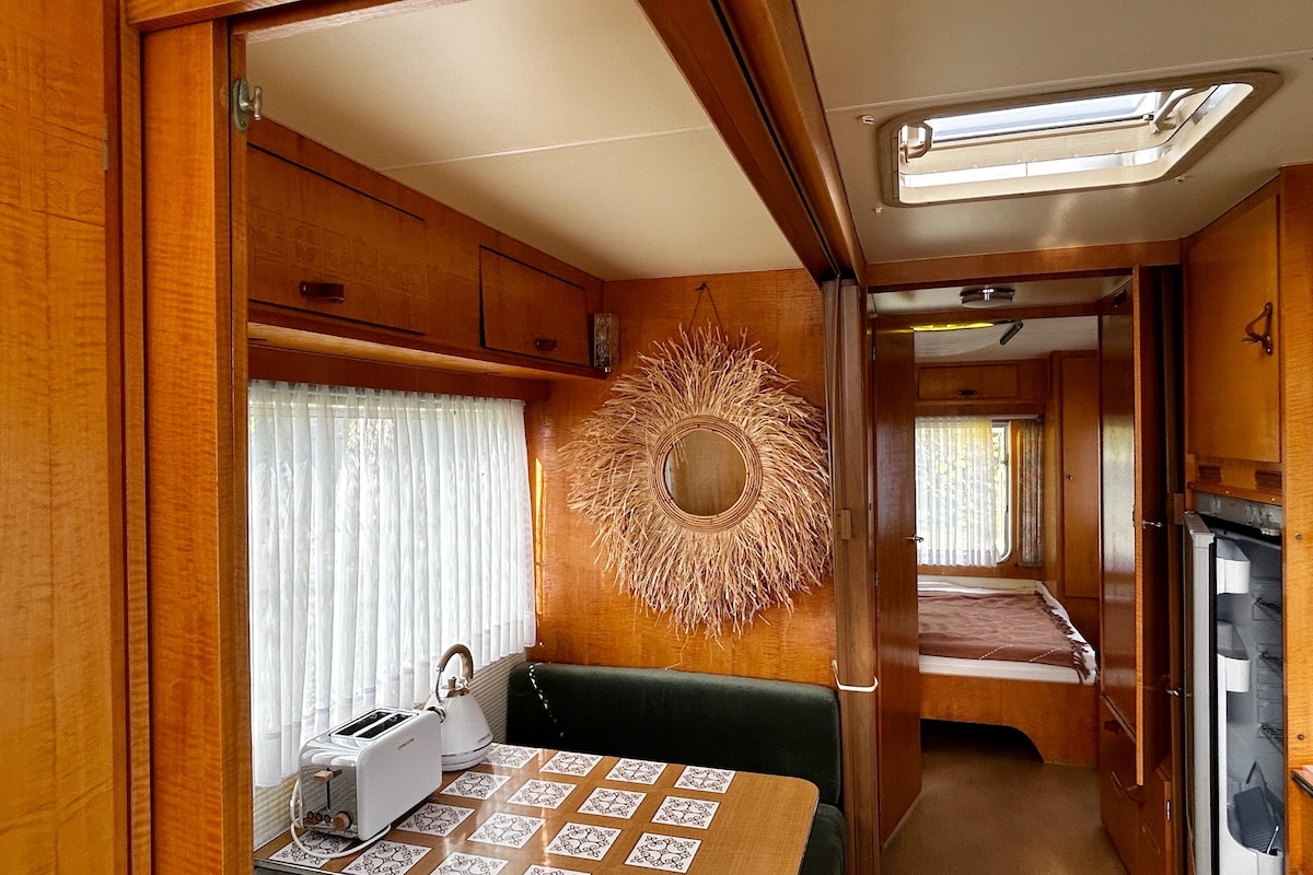 The interior of the vintage Tabbert caravan features warm wooden paneling and a cozy seating area with a table. A decorative mirror and natural elements enhance the inviting feel. Natural light filters through sheer curtains, illuminating the space and creating a comfortable atmosphere.
