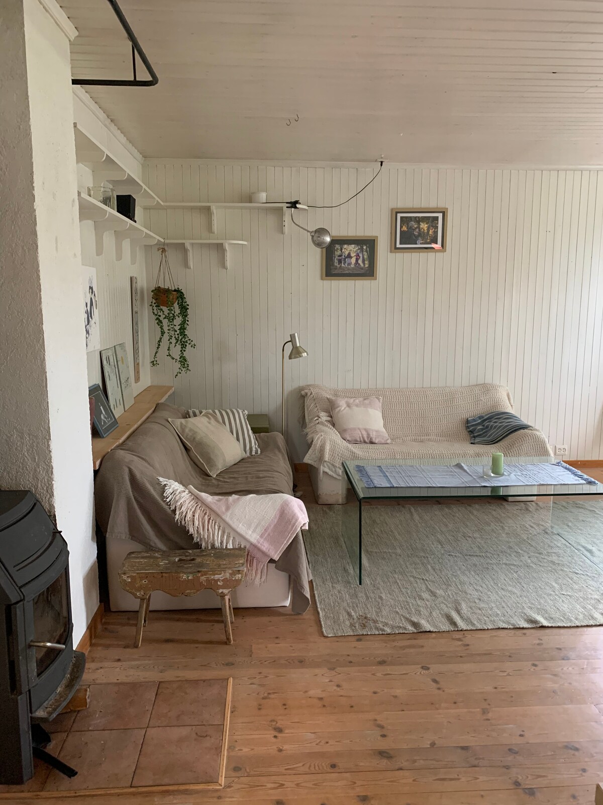A cozy living area features two soft sofas adorned with blankets, a glass coffee table, and a light green candle. Wooden flooring is complemented by a large rug, while artwork hangs on the white paneled walls, creating a calm and relaxed space.