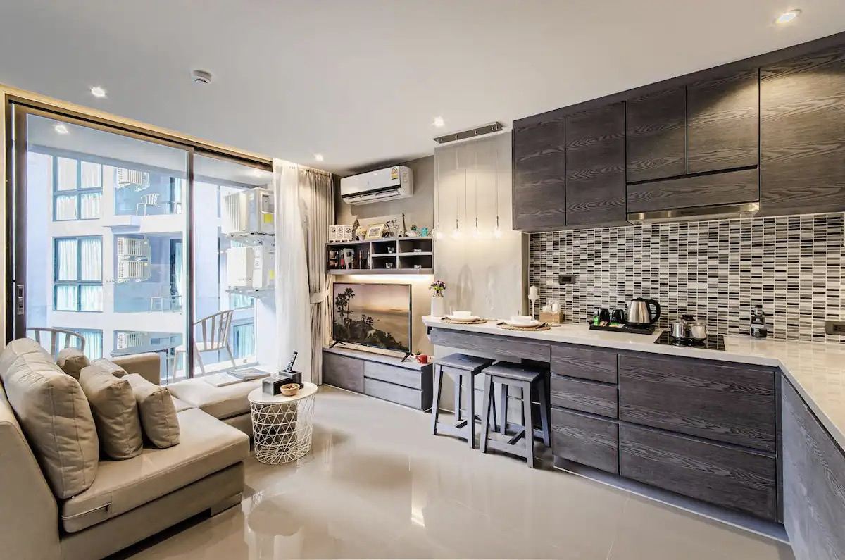 An open-concept kitchen and living area is equipped with modern appliances and a stylish backsplash. Seating is provided by a cozy sofa and bar stools. Large glass doors lead to a balcony, allowing natural light to fill the space.