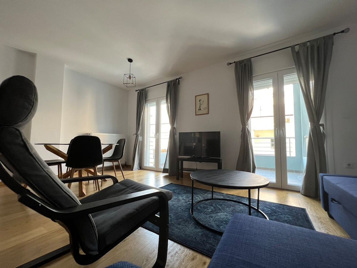 A modern living area is highlighted by large windows draped with soft gray curtains, allowing natural light to fill the space. A round table with four chairs is positioned nearby, while a cozy sofa and an armchair offer comfortable seating around a low coffee table.
