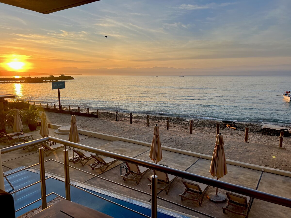 An expansive balcony offers unobstructed views of a serene sunrise over the ocean. Lounge chairs are arranged around a pool, with umbrellas providing shade. The beachfront is visible below, alongside calm water reflecting orange and pink hues from the sky.