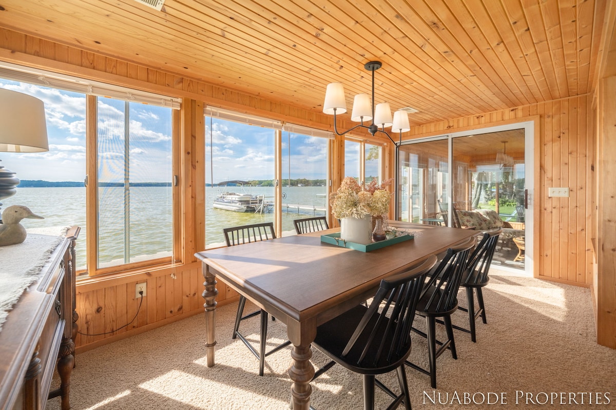A sunlit dining area is framed by large windows that offer scenic views of Hess Lake. The wood-paneled walls and ceiling create a natural feel. A wooden dining table is surrounded by six black chairs, inviting guests to gather for meals and conversation.