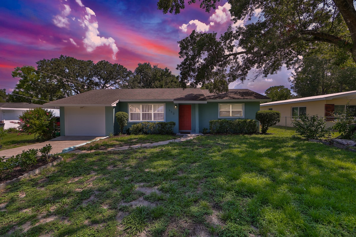 A cozy home with a light blue exterior is surrounded by lush green grass and landscaping. A welcoming red door is prominently featured at the front. The sky showcases a vibrant blend of colors, adding to the tranquil ambiance of the property.