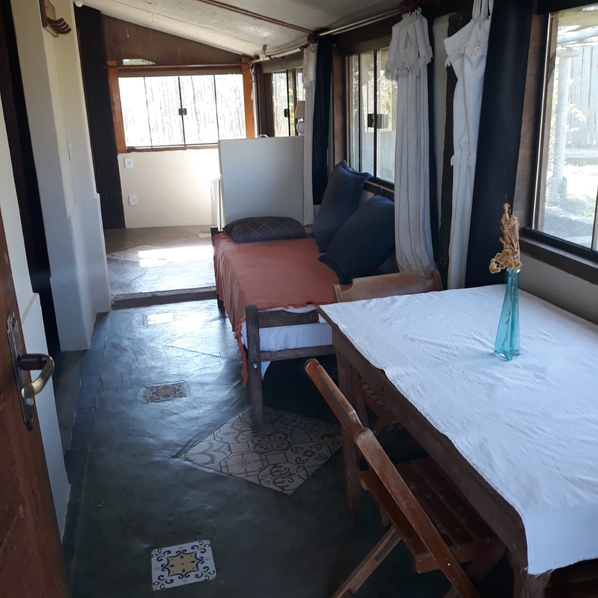 The interior space features a bright layout with a sofa bed and a table. Sunlight streams through large windows, and nature is visible outside. The floor showcases a mix of patterned tiles, contributing to the overall rustic charm of the area.