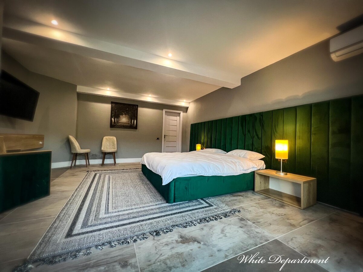 A spacious room features a large bed with a green upholstered headboard and white linens. Two light-colored chairs are positioned beside the bed. Soft lighting is provided by a lamp on a wooden nightstand, enhancing the neutral color scheme and simple decor.