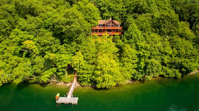 A spacious wooden house is nestled among lush greenery, with a long dock extending into the calm waters of Lake Nantahala. The surrounding trees create a natural backdrop, highlighting the serene setting aimed at relaxation and outdoor activities.