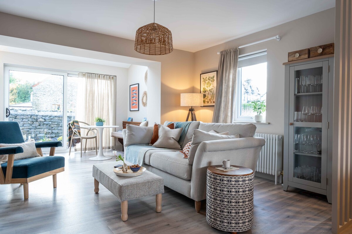 An open plan living area displays modern furnishings, including a comfortable sofa and accent chairs. Natural light filters through large windows, enhancing the soft, neutral color palette. A coffee table, adorned with decorative items, complements a side table with a cup. A dining area is subtly visible.