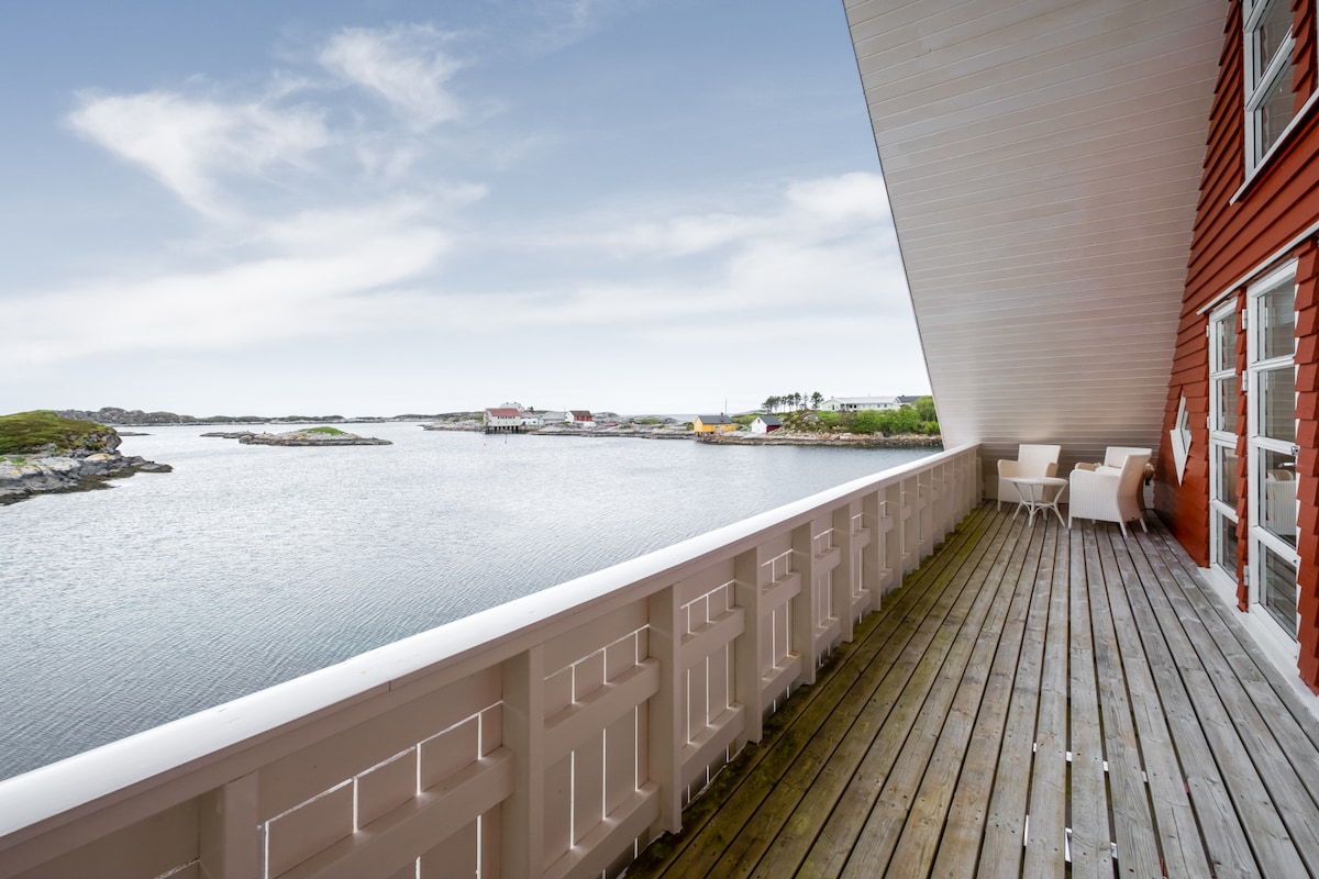 A spacious balcony features wooden flooring and a protective railing. White outdoor chairs invite relaxation, while scenic views of calm waters and distant islets are visible. The image captures an open sky, enhancing the connection between indoor and outdoor living.