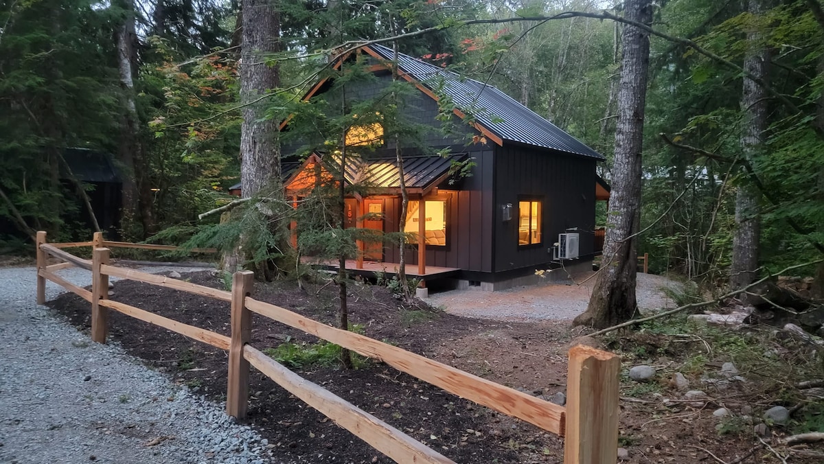 The Bearfoot Chalet is nestled among tall trees, featuring a modern black exterior and large windows illuminated warmly from within, inviting nature indoors. A gravel path leads to the home, which is bordered by a wooden fence, enhancing its secluded mountain setting.