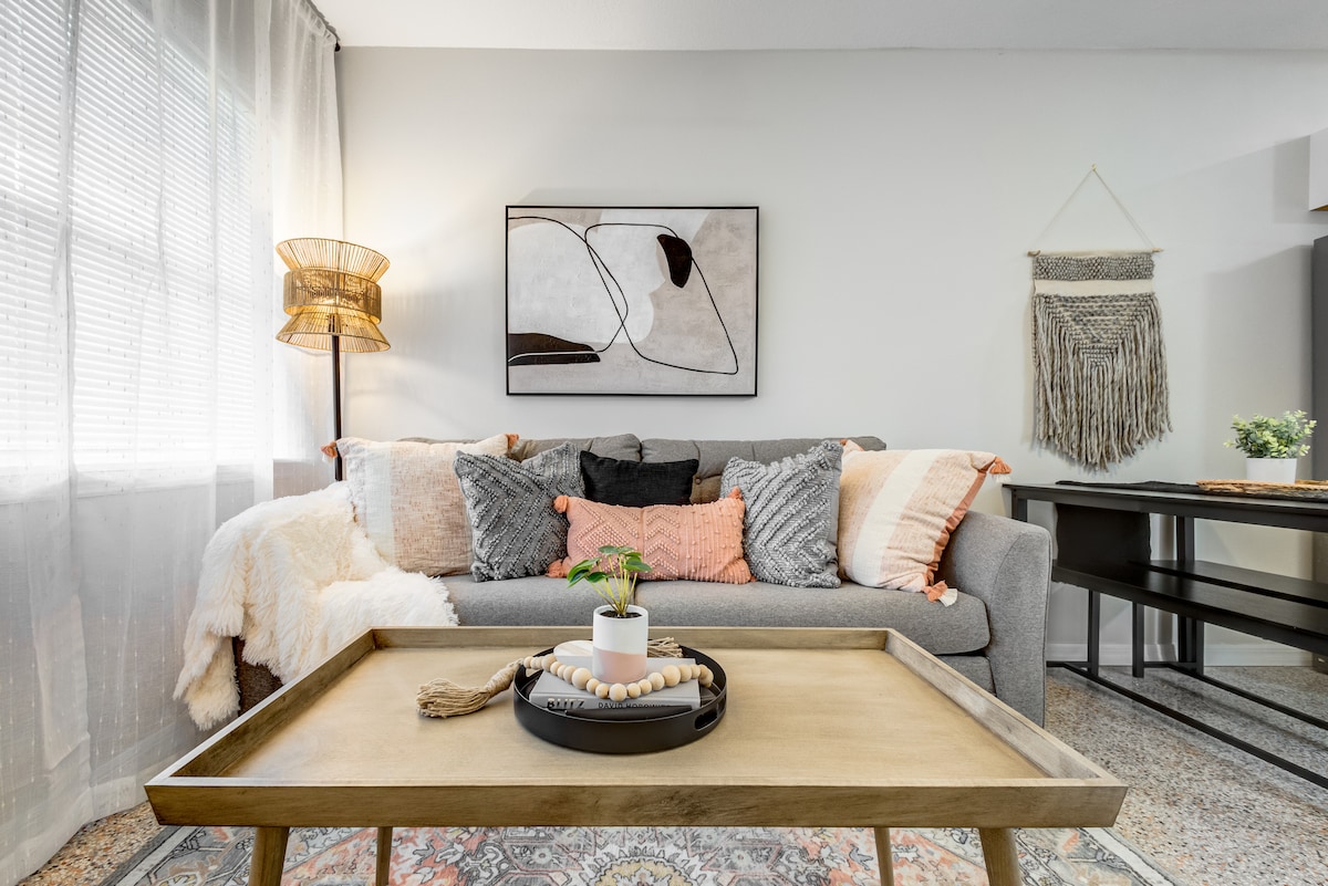 A modern living area is displayed with a comfortable sofa adorned with a mix of textured cushions in various colors. A wooden coffee table sits in the center, featuring a small potted plant and decorative items. Natural light filters through sheer curtains, creating a soft ambience.