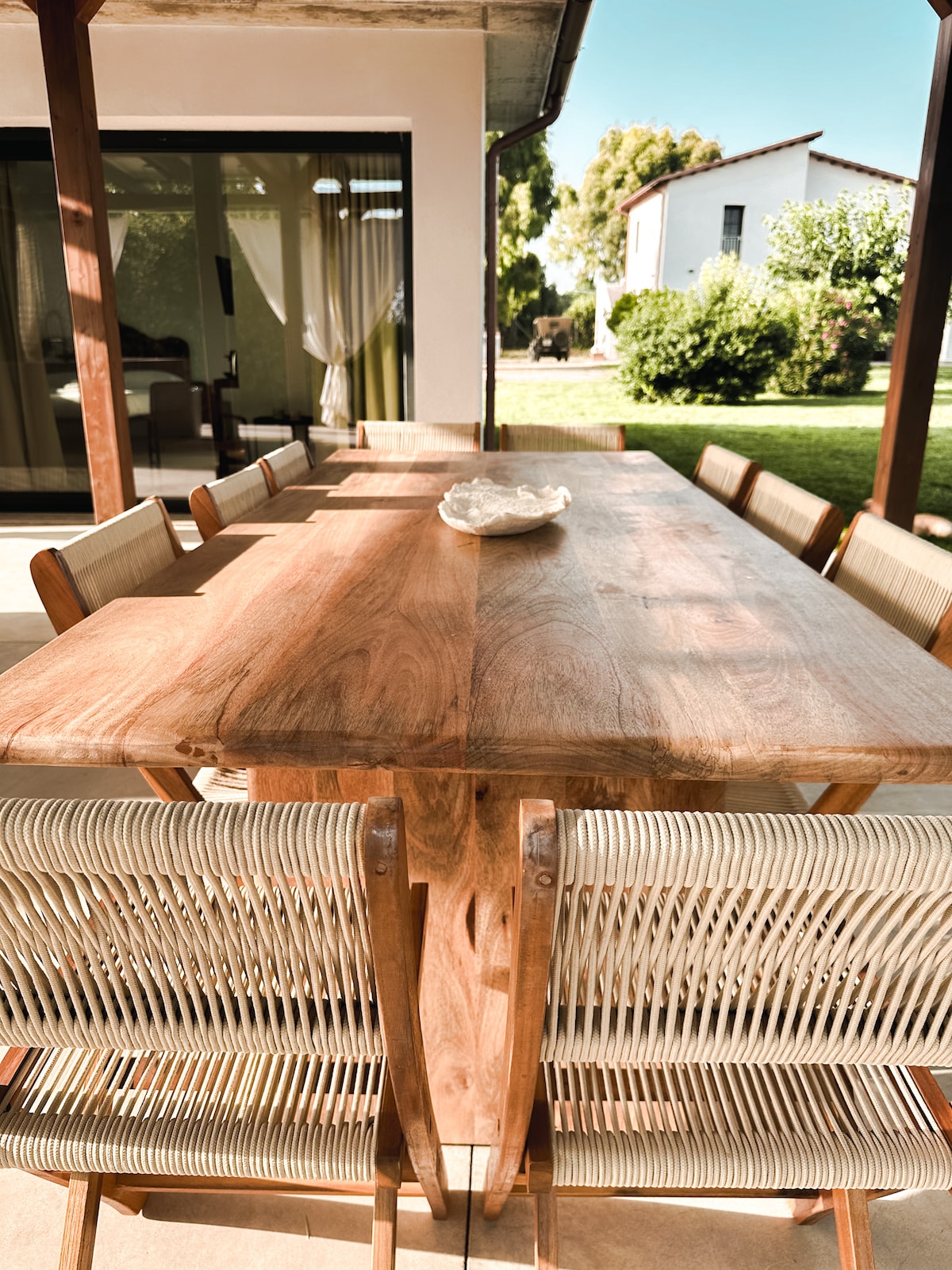 A large wooden dining table with eight woven chairs is set on a patio area. A decorative bowl is placed at the center. The background features a lush garden, with well-maintained grass and trees visible in the distance, enhancing the outdoor dining experience.