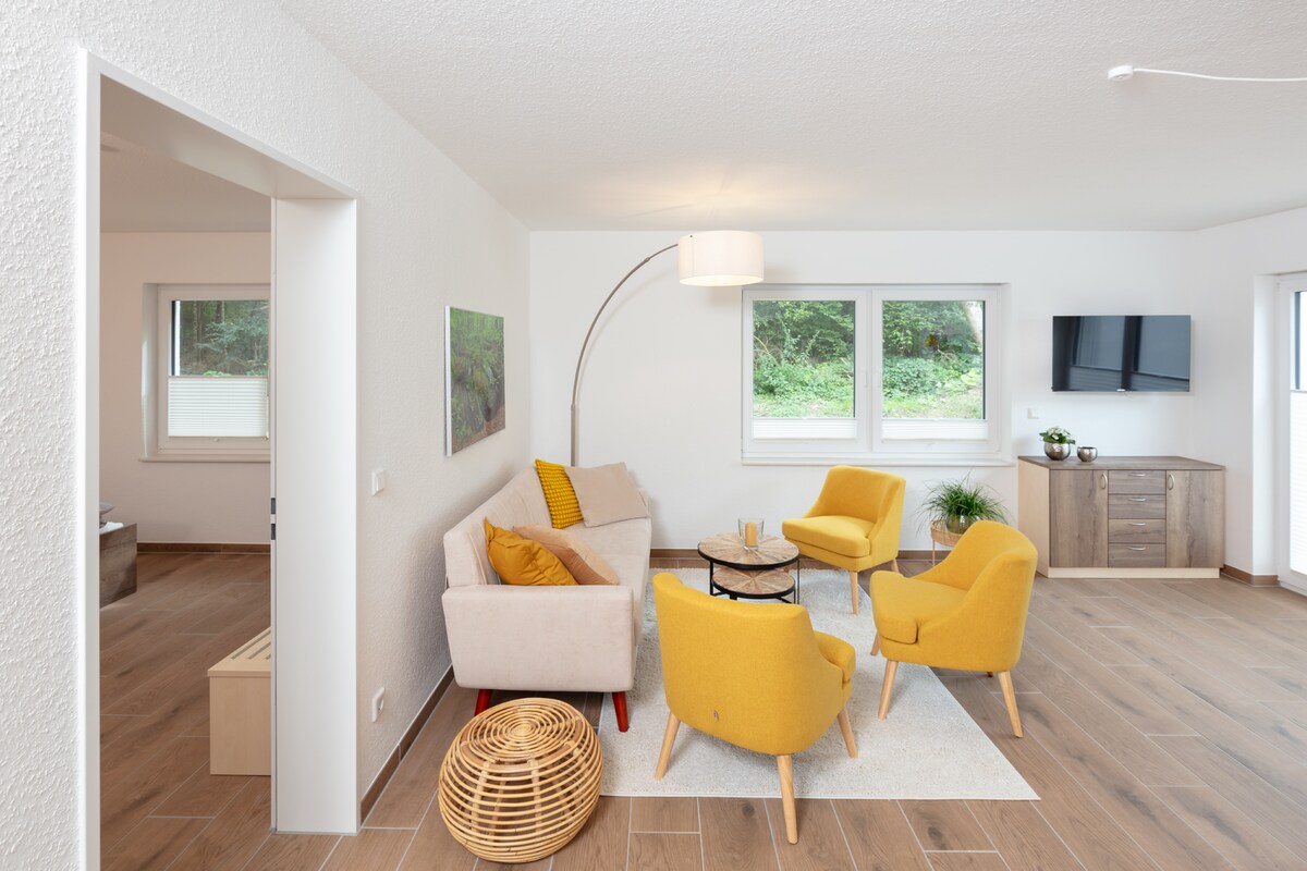 The living area features a comfortable seating arrangement with a light-colored sofa and two yellow accent chairs around a small table. A modern floor lamp adds soft lighting, and a flat-screen television is mounted on the wall, enhancing the room's functionality.