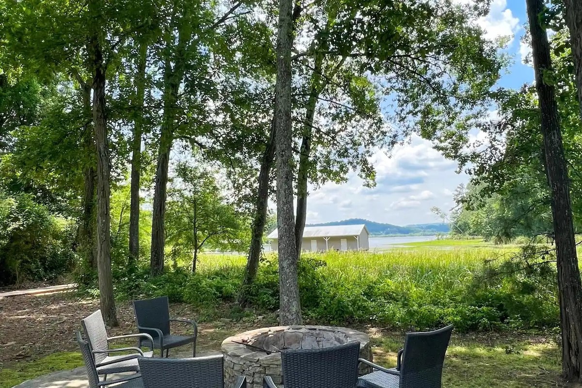 A serene outdoor area is surrounded by trees, with a stone fire pit at the center. Black chairs are arranged around the fire pit, offering a cozy space to relax. In the background, a private boathouse is visible, set against a backdrop of green grass and the lake.
