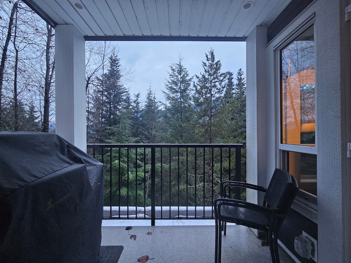 A covered outdoor balcony is visible, featuring a black grill and a chair. The view highlights tall evergreen trees, contributing to a serene atmosphere. The balcony is bordered by black railings, with a portion of the building's exterior also shown.