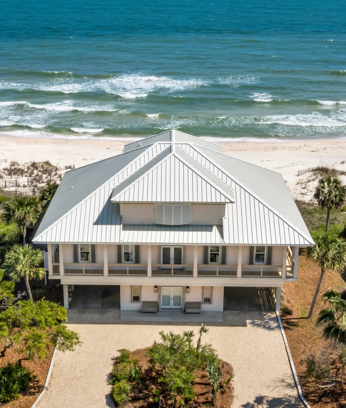 An aerial view of a beachfront home reveals a charming architecture with a metal roof. The house is framed by palm trees and overlooks the sandy shore and waves of the Gulf of Mexico. A spacious driveway leads to the entrance.