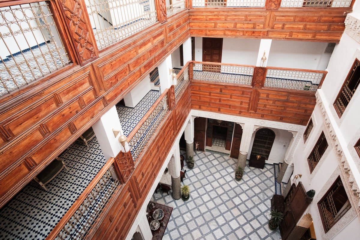 An aerial view of the central courtyard features a geometric tile pattern, surrounded by traditional archways and wooden balconies. Potted plants are positioned throughout, enhancing the space's tranquility. Natural light fills the area, highlighting the craftsmanship of the wooden details and the overall design.