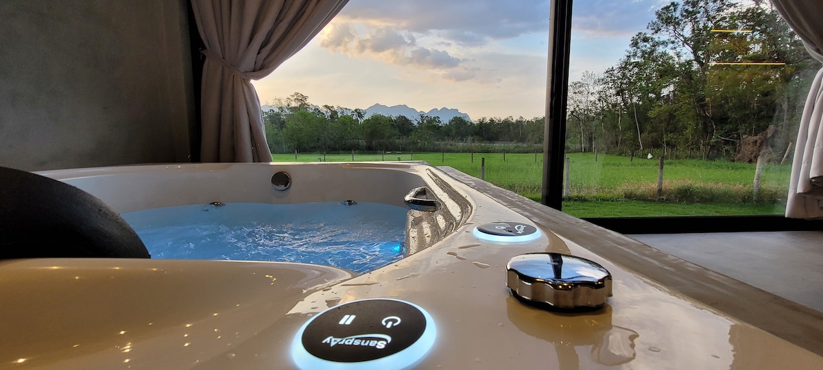 A spacious hot tub is positioned by a large window, offering views of the scenic landscape and distant mountains. Natural light filters through sheer curtains, illuminating the water's surface and surrounding area, creating a serene atmosphere.