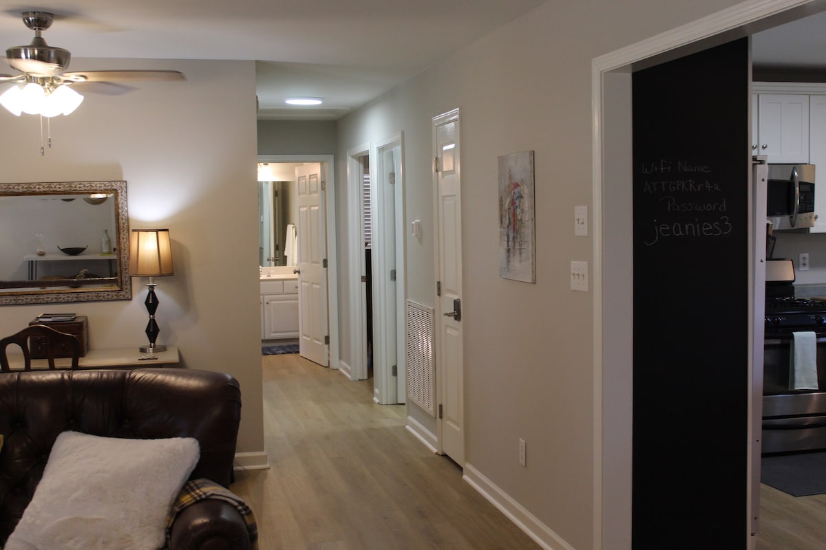 A well-lit living area features a leather sofa and a stylish lamp. A hallway is visible, leading to a bathroom and additional rooms. A chalkboard on the wall displays WiFi details, while art adorns the space, contributing to a comfortable ambiance.