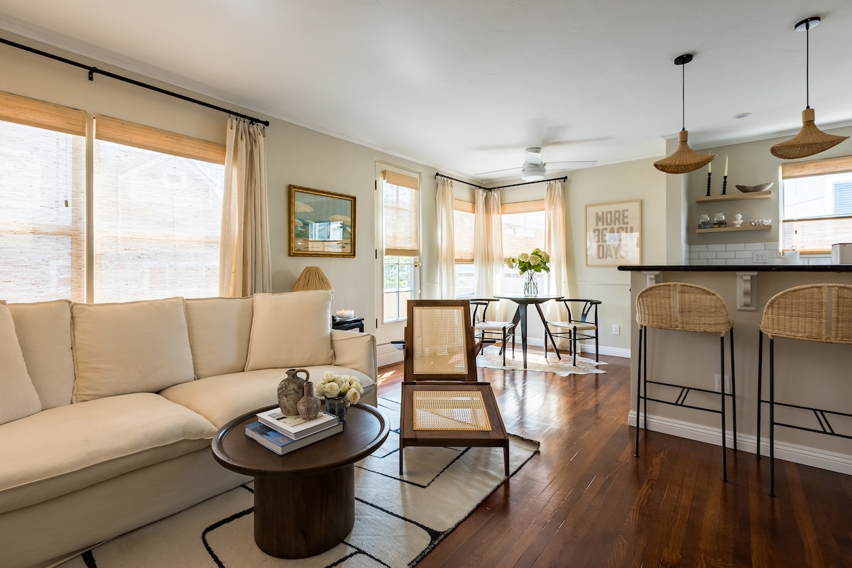 The open living space features a light-colored sofa with plush cushions, accompanied by a circular wooden coffee table. Large windows allow natural light to fill the room, enhancing the comfortable ambiance. A bar area with two stylish stools is visible in the background.