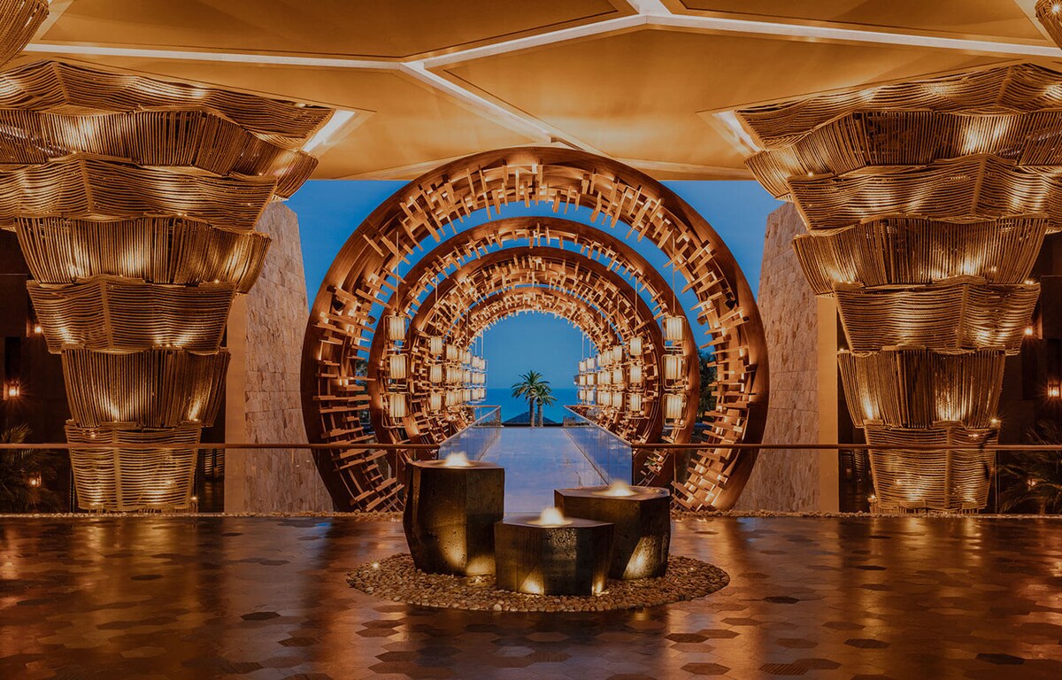 An elegant entrance is framed by a circular, illuminated tunnel design, featuring intricate metallic elements. Soft lighting highlights the structure's contours, while a serene pool reflects the evening sky. Potted greenery adds a touch of nature to the welcoming ambiance.