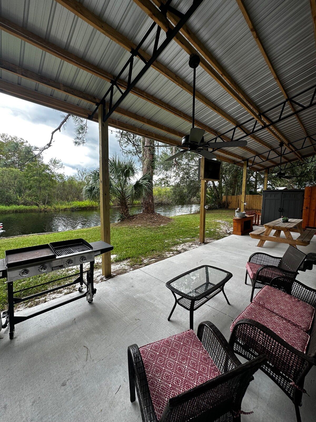 A covered patio area features a barbeque grill and a small glass table surrounded by two chairs with cozy cushions. A picnic table is visible nearby, with a serene view of the canal and lush greenery in the background, enhancing the outdoor ambiance.