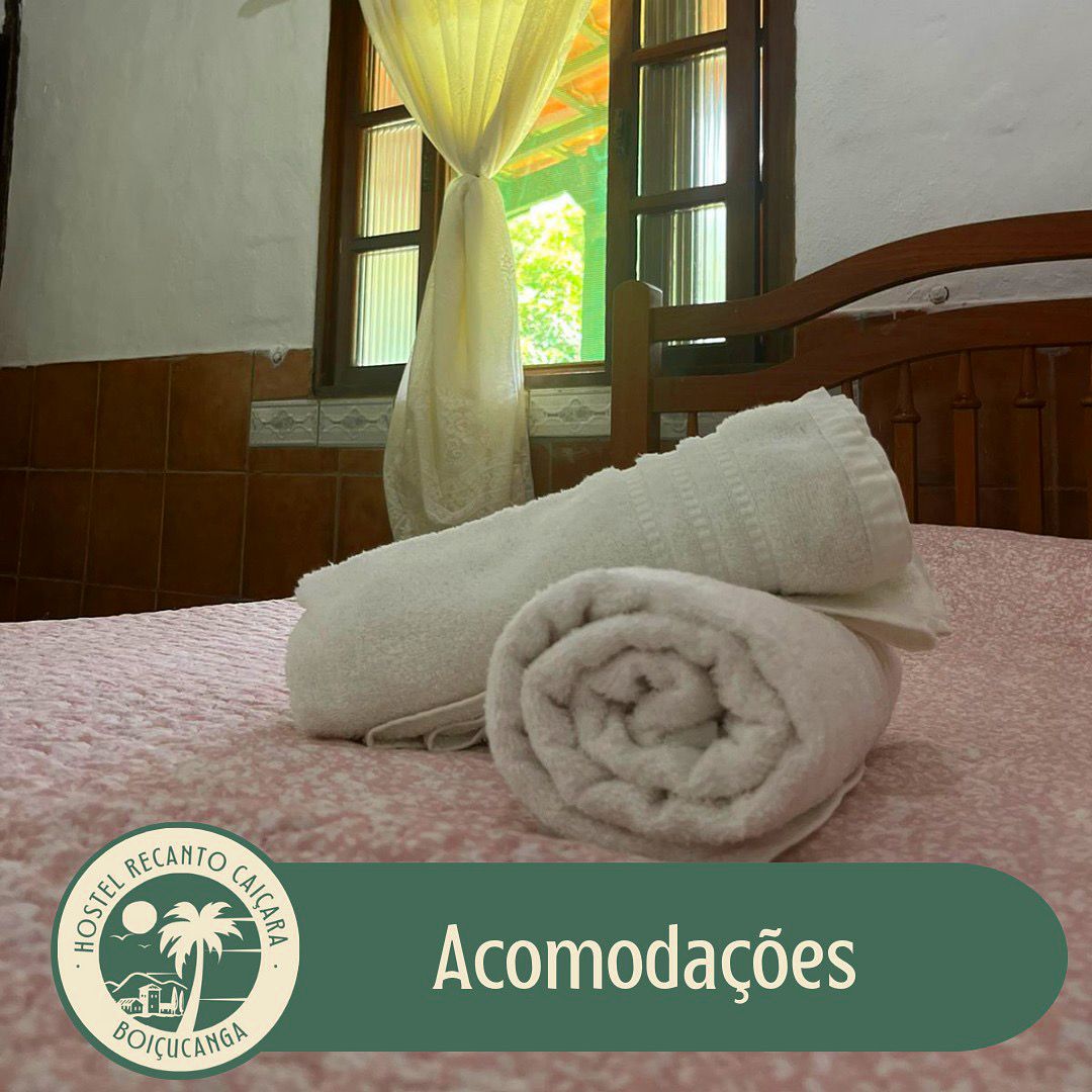 A cozy sleeping area features a neatly made bed topped with a light pink bedspread. Rolled towels are placed on the bed, with soft natural light filtering through the window adorned with light-colored curtains.
