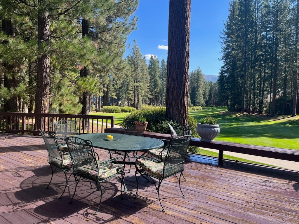 A spacious deck is shown with a round table surrounded by four metal chairs featuring floral cushions. Tall pine trees line the edge of the deck, overlooking the landscaped golf course, with a clear blue sky visible in the background.