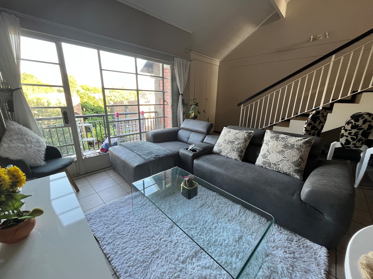 A spacious living area features a dark sectional sofa adorned with decorative pillows, positioned on a plush white rug. Large glass sliding doors offer access to a balcony, filling the space with natural light. A coffee table and potted plant provide additional elements to the room.