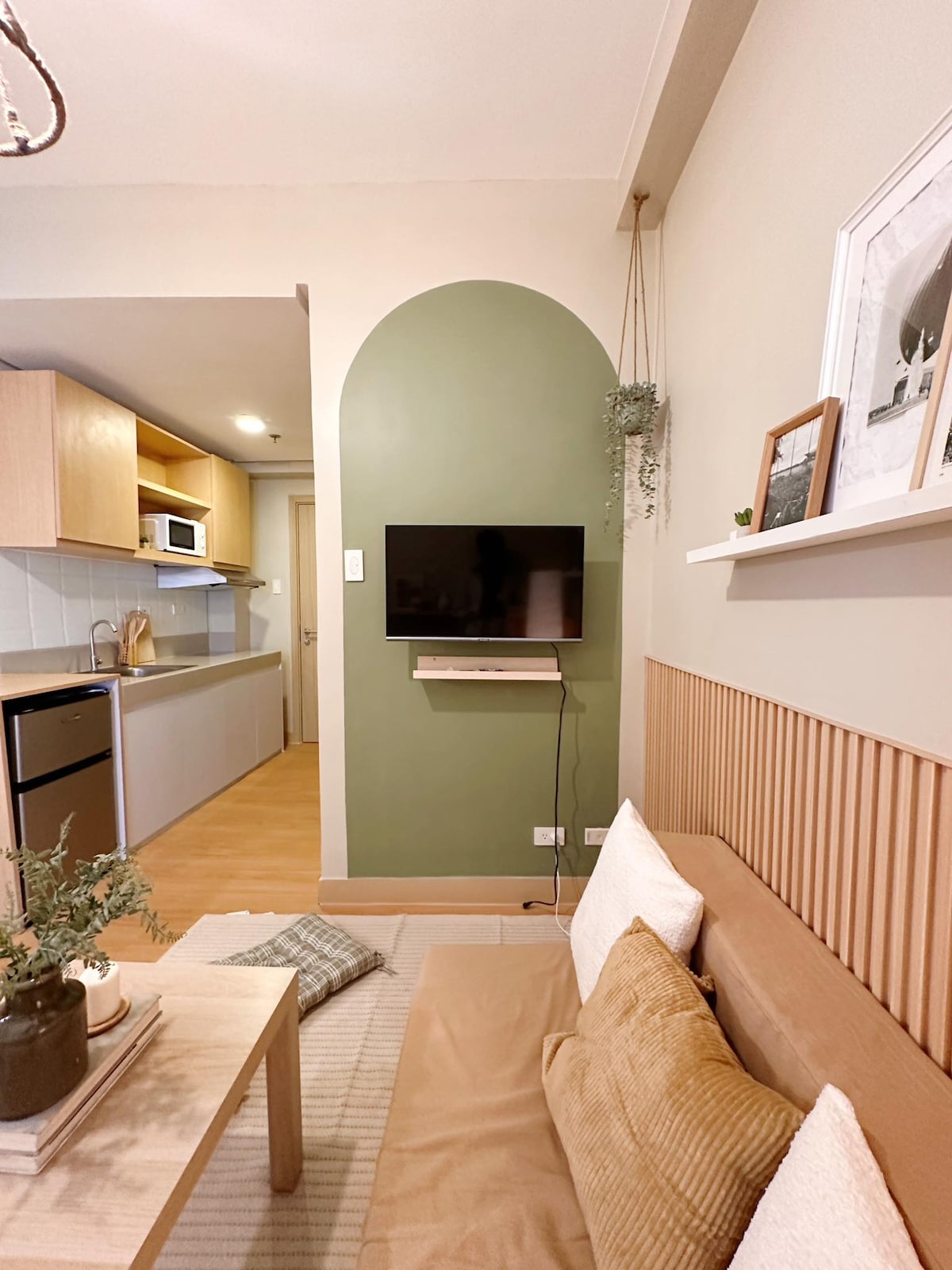 A living area features a light brown sofa adorned with cushions, placed against a soft green wall. A minimalistic wooden shelf displays framed art, while a television is mounted above a small wooden table. The kitchenette is visible in the background, showcasing modern appliances.