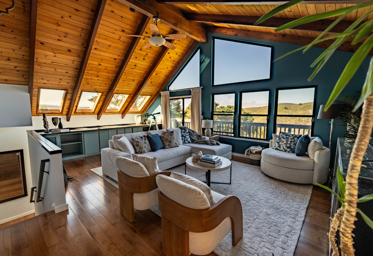 A spacious living area features a high A-frame ceiling with exposed wooden beams and several triangular windows. Comfortable seating arrangements include plush sofas and armchairs, arranged around a central coffee table. Sunlight filters through the windows, highlighting the modern decor and offering views of the surrounding landscape.