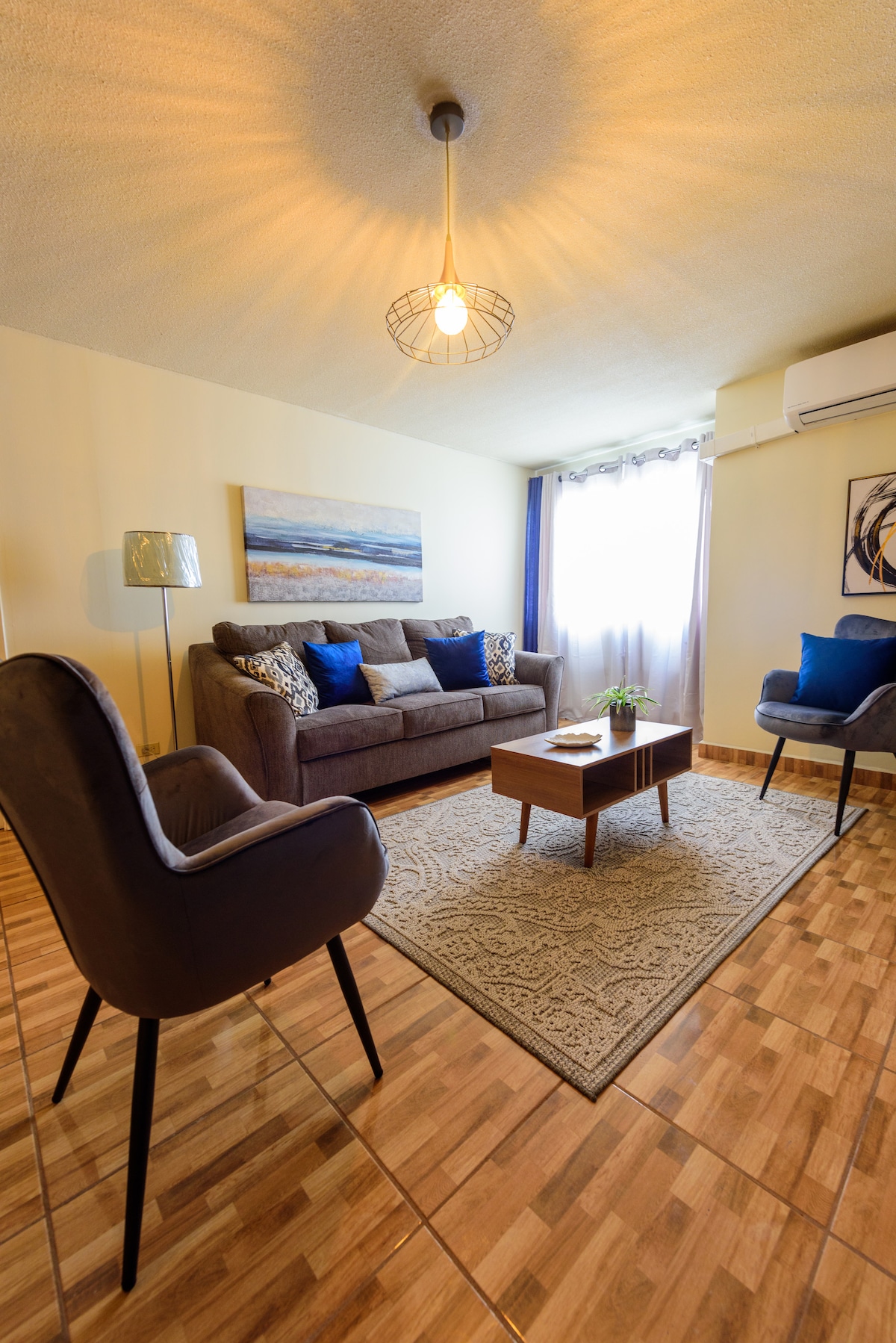 A well-lit living area is furnished with a comfortable sofa, two accent chairs, and a coffee table. A decorative rug lies on the floor, with light filtering through a window draped with sheer curtains. Artwork featuring calming tones adorns the wall.