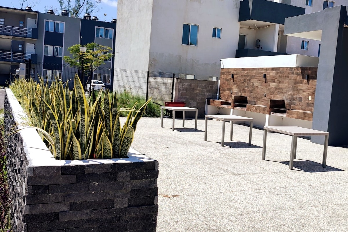 A landscaped outdoor area features planters filled with tall green plants, complemented by several white tables arranged throughout the space. A stone wall with a textured finish adds depth, while modern buildings are visible in the background.