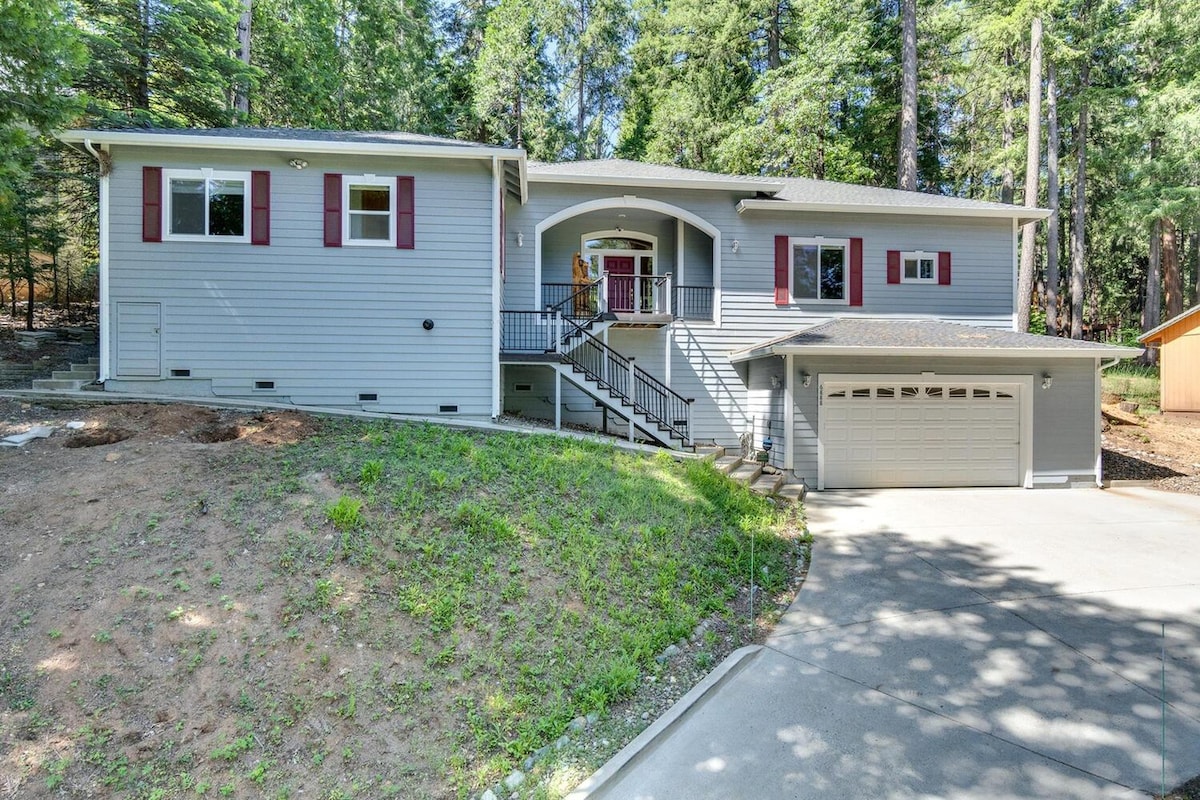 The modern house is surrounded by tall trees, featuring a light gray exterior with red shutters. A gently sloped driveway leads to a spacious garage. Steps with railings provide access to an inviting front porch, enhancing the home's welcoming entrance.