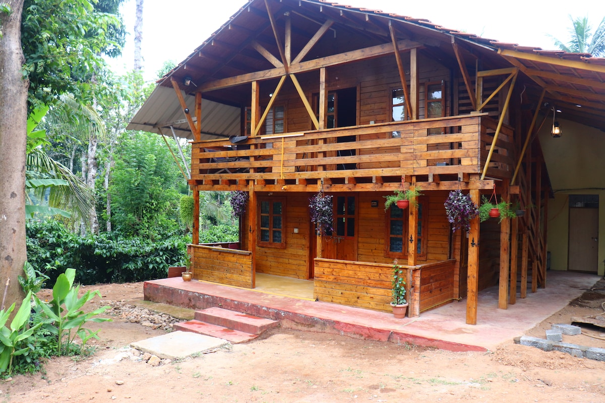 A rustic wooden villa is showcased, featuring a spacious porch supported by wooden beams. Flower pots adorn the balcony, and the exterior is surrounded by lush greenery, enhancing the natural setting. A pathway leads to the entrance, inviting guests to explore the serene environment.