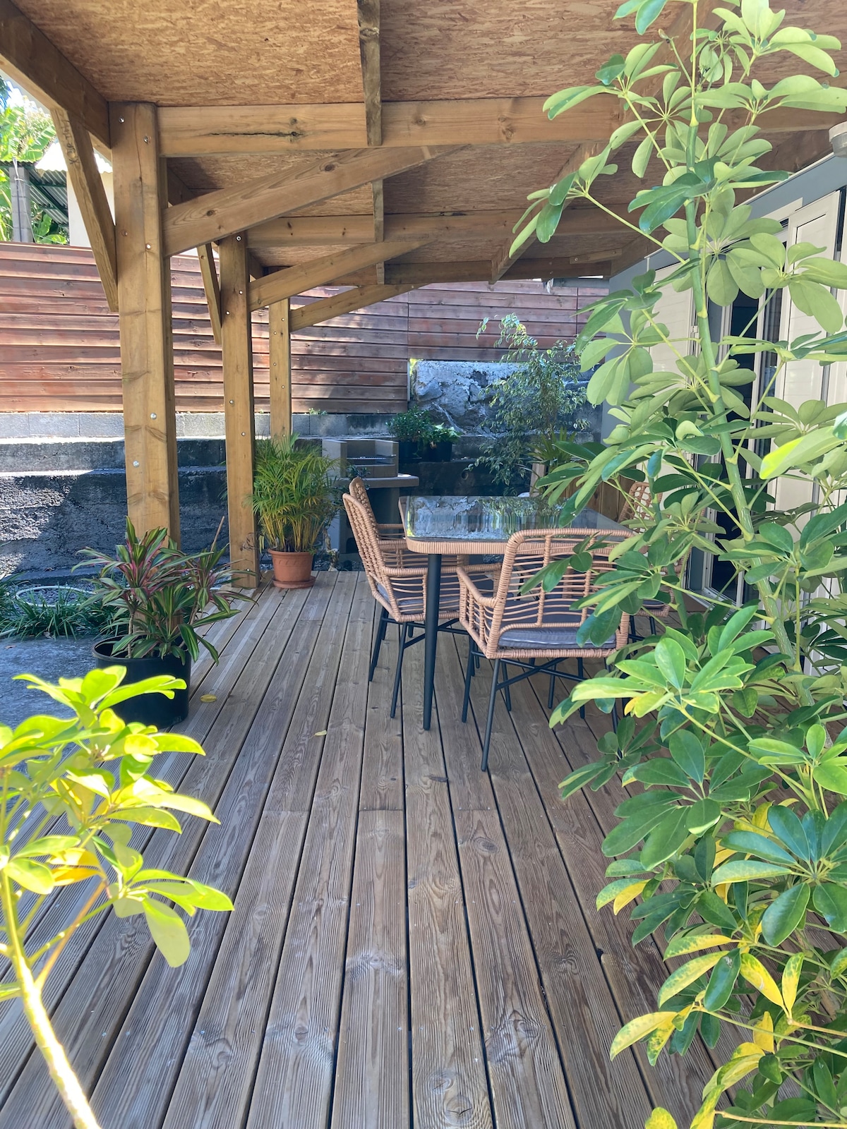 An outdoor seating area features two rattan chairs with a small table, surrounded by greenery. The wooden deck adds warmth, and the structure is covered by a wooden overhang, providing shade. Lush plants in various pots enhance the welcoming environment.