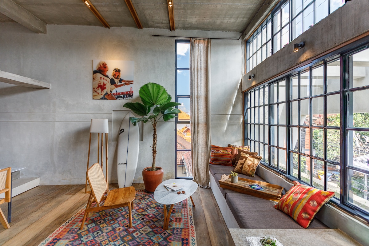 The loft features large windows allowing abundant natural light to illuminate the space. A comfortable seating area includes a stylish sofa adorned with colorful cushions and a low coffee table. A decorative plant adds a touch of greenery, while artwork enhances the modern aesthetic.