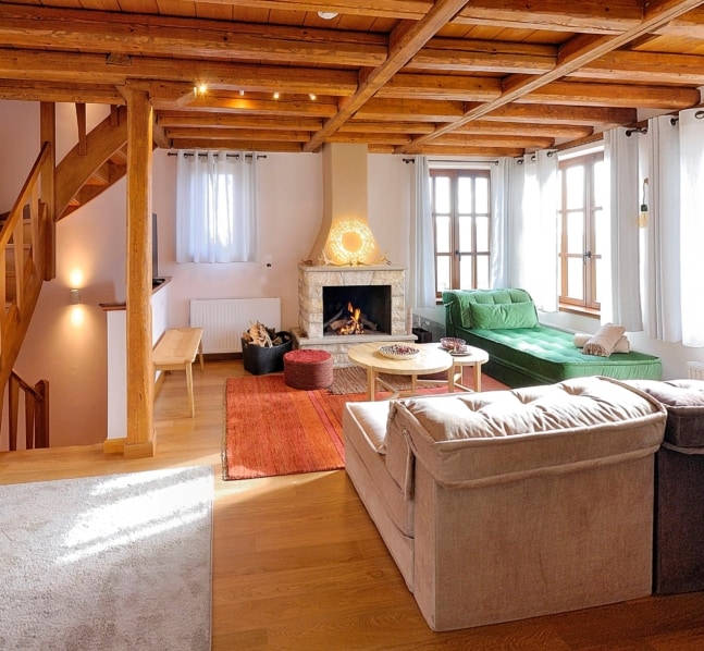 A spacious living area is highlighted by a stone fireplace, offering a cozy setting. Soft seating arrangements include a green sofa and a light-colored couch. Large windows allow natural light to fill the space, complemented by wooden beams and a warm wood floor.