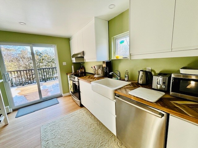 A modern kitchen is shown, featuring sleek white cabinetry and a spacious sink. Light wood countertops and stainless-steel appliances are visible. A sliding glass door leads to a deck, allowing natural light to brighten the space while a light-colored rug rests beneath the counter area.