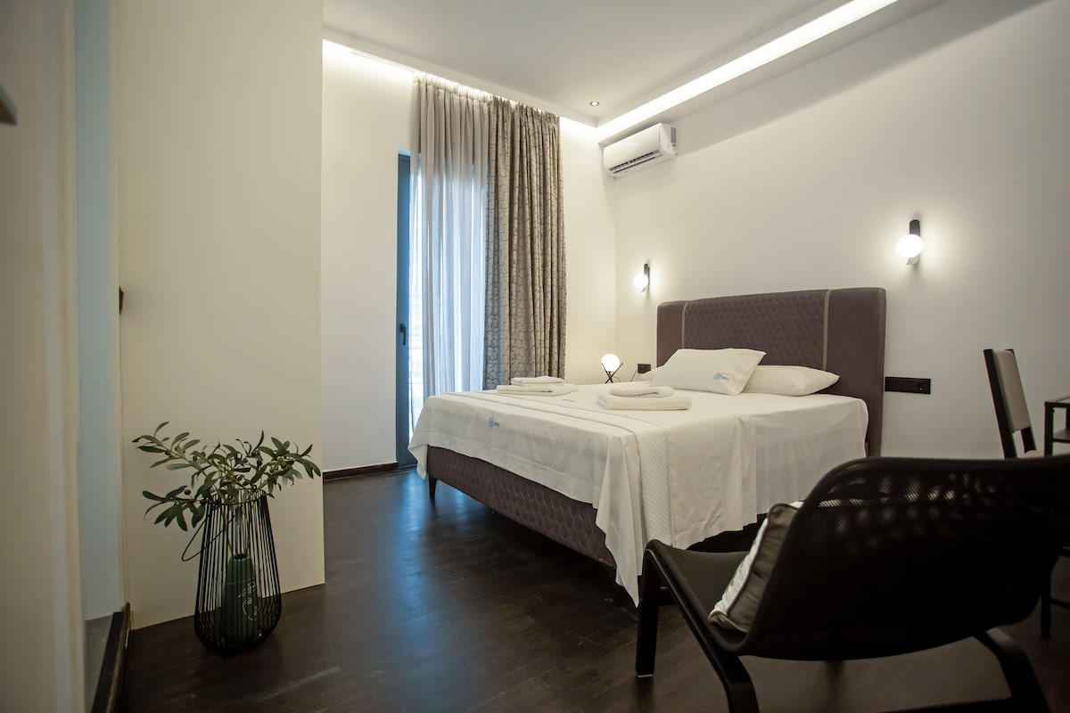 A modern bedroom features a spacious bed with crisp white linens, complemented by a soft headboard. Large windows are adorned with sheer curtains, allowing natural light to filter in. A small plant in a decorative pot adds a touch of greenery, while minimalist furniture enhances the room's clean design.