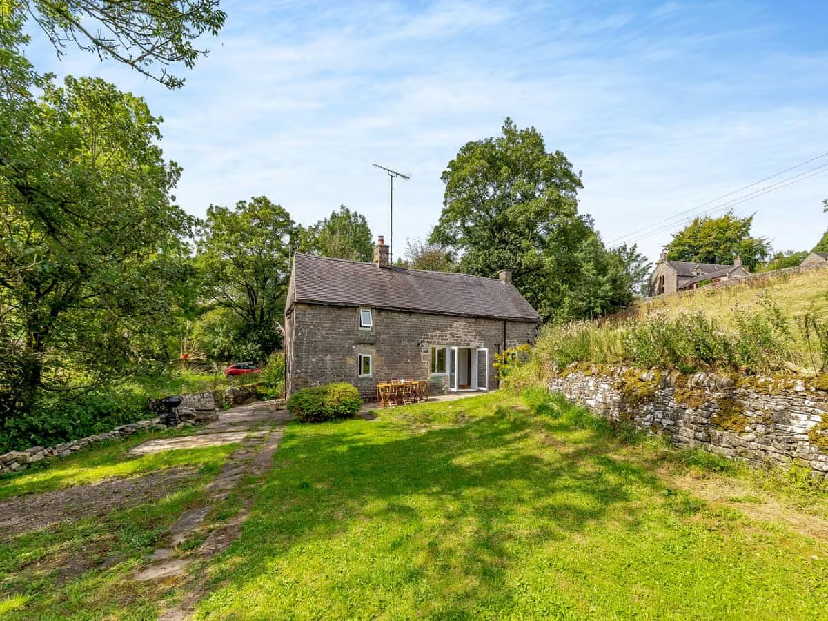 A historic stone cottage is set amidst greenery, showcasing a well-tended garden with large areas of lawn. The patio features outdoor seating, surrounded by natural stone walls. The entrance is marked by large patio doors, leading into the welcoming interior.