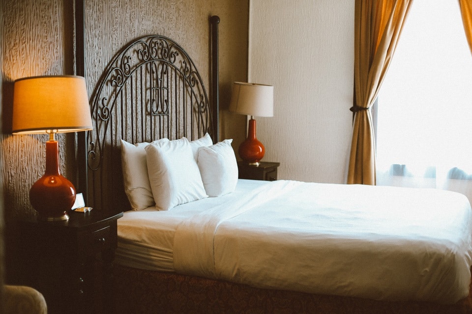 A king-size bed with white bedding is framed by an intricately designed headboard. Soft lighting is provided by two bedside lamps with orange bases. Light filters in through sheer curtains, creating a warm ambiance in the room.