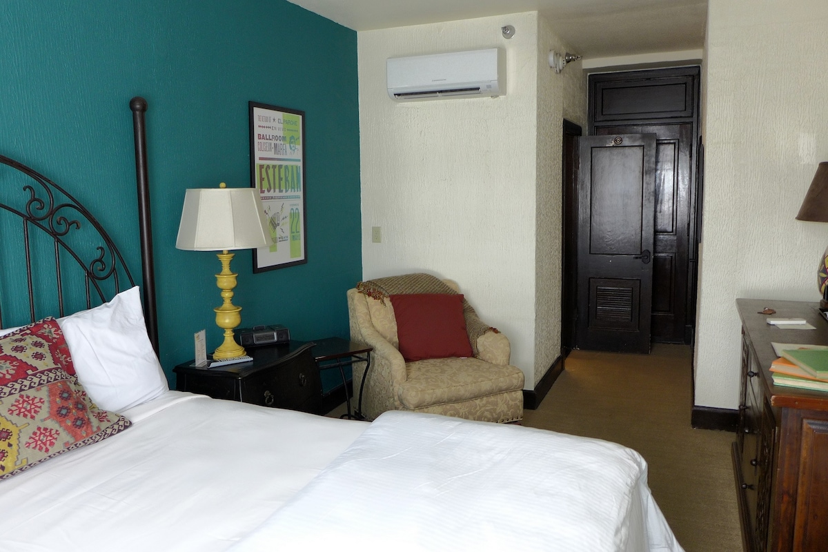 A guest room showcases a spacious bed with a white duvet, complemented by decorative pillows. A cozy armchair sits in the corner, paired with a bedside table featuring a yellow lamp. The wall behind the bed is painted in a soothing teal, enhancing the room's inviting ambiance.