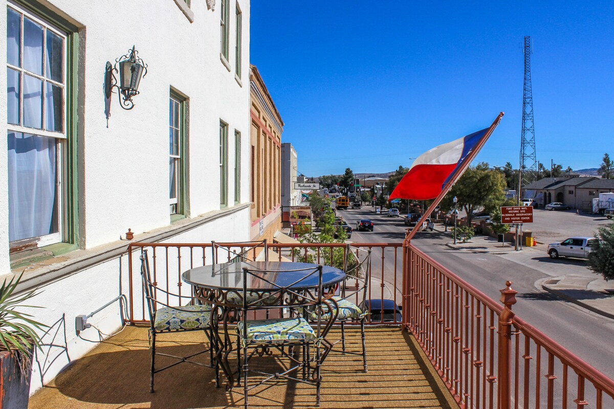 A private balcony features a round table surrounded by wrought iron chairs with colorful cushions. The Texas flag is displayed, fluttering in the breeze. A view of the bustling street below is visible, framed by historic buildings and clear blue skies.